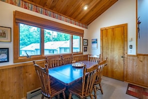 Uma sala de jantar com uma mesa e cadeiras de madeira, janelas amplas com vista para casas, e uma porta de madeira.