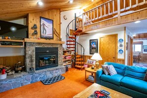 A cozy living room with a stone fireplace, a blue sofa, a wooden staircase, and a television on the wall.