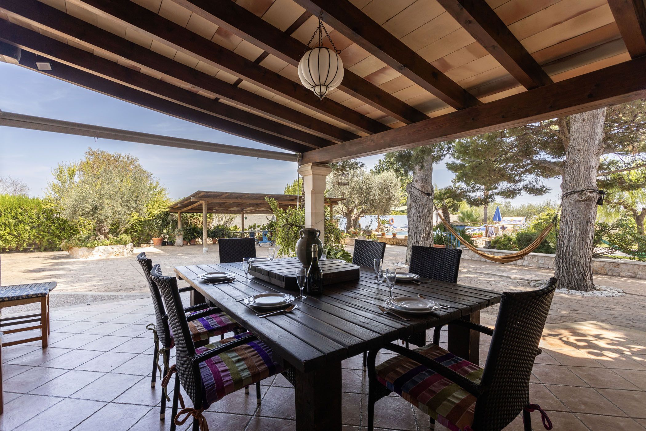 An outdoor covered patio with a wooden table set for dining, surrounded by wicker chairs, and a hammock.