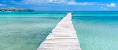 A wooden pier extending into clear turquoise water, leading to a distant landmass with mountains.