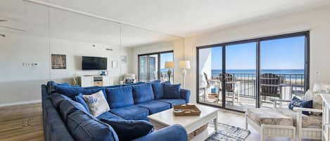 A modern living room with a blue sectional sofa, a flat-screen TV, and a view of the ocean through large windows.