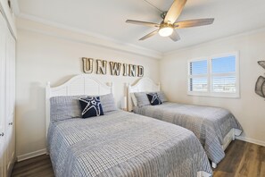 A bedroom with two beds, a ceiling fan, a window with blinds, and a wall with the word 'UNWIND' spelled out.