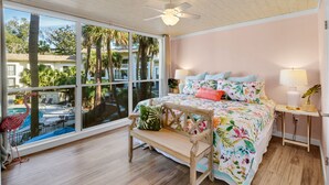 Une chambre avec une grande fenêtre, un ventilateur de plafond, un lit avec une literie à motifs floraux, un banc et une table de chevet avec une lampe.