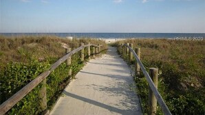 Un sentier sablonneux menant à la plage, bordé de rambardes en bois et de végétation.