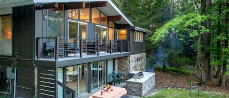 A modern two-story house with a balcony, a hot tub, and a patio with a table and chairs.