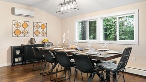 Uma sala de jantar com uma mesa comprida posta para seis pessoas, cadeiras pretas, um candelabro e uma janela com vista para árvores.