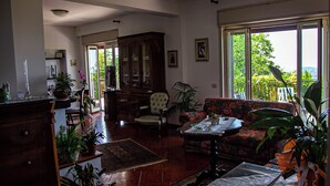 A living room with a sofa, armchair, and a dining table with a laptop. There are potted plants and a cabinet.