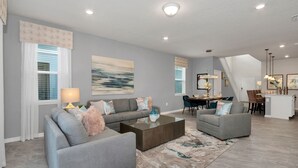 A modern living room with a gray sofa set, a coffee table, and a dining area with a chandelier. - Newly Built 6BR Home w/Private Pool, 15min to Disney! *FREE Resort/ Water Park* (Davenport)