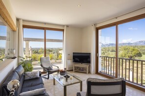 A spacious living room with a sofa, armchair, and a glass coffee table. A flat-screen TV is on a stand, and the room has large windows offering a view of the countryside. - Modern villa with 3 bedrooms, 2 bathrooms, wifi, terraces and pool (Inca)