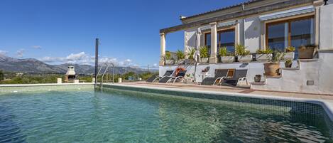 A clear swimming pool with lounge chairs and potted plants on a terrace, overlooking a mountainous landscape.