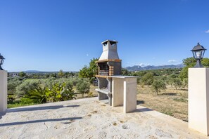Un patio de piedra con una estructura de chimenea y un farol, con vistas a un paisaje rural con árboles y montañas distantes.