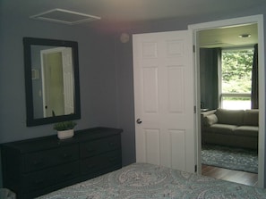 A bedroom with a dark dresser, a mirror, a white door, and a view of a living room through an open door.