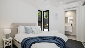 A modern bedroom with a large bed, a bedside table with a lamp, and a view of the outside through a glass door. - Husky Sands - Pet Friendly - 5 Mins Walk to Beach (Huskisson)