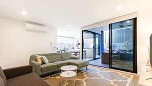 A modern living room with a grey sofa, a round coffee table, and a patterned rug. There is a sliding glass door leading to a balcony with a view of the outside. - Husky Sands - Pet Friendly - 5 Mins Walk to Beach (Huskisson)
