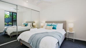 Een moderne slaapkamer met een grote spiegel, een bed met witte lakens, een grijs hoofdeinde en een klein nachtkastje met een lamp.