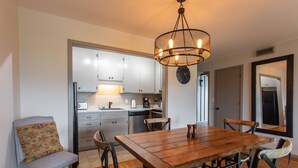 Un comedor con mesa y sillas de madera, una araña y una cocina al fondo.