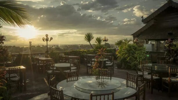 An outdoor patio with round tables and chairs, surrounded by lush greenery and a view of the cityscape during sunset. - Pakse Hotel (Pakse)