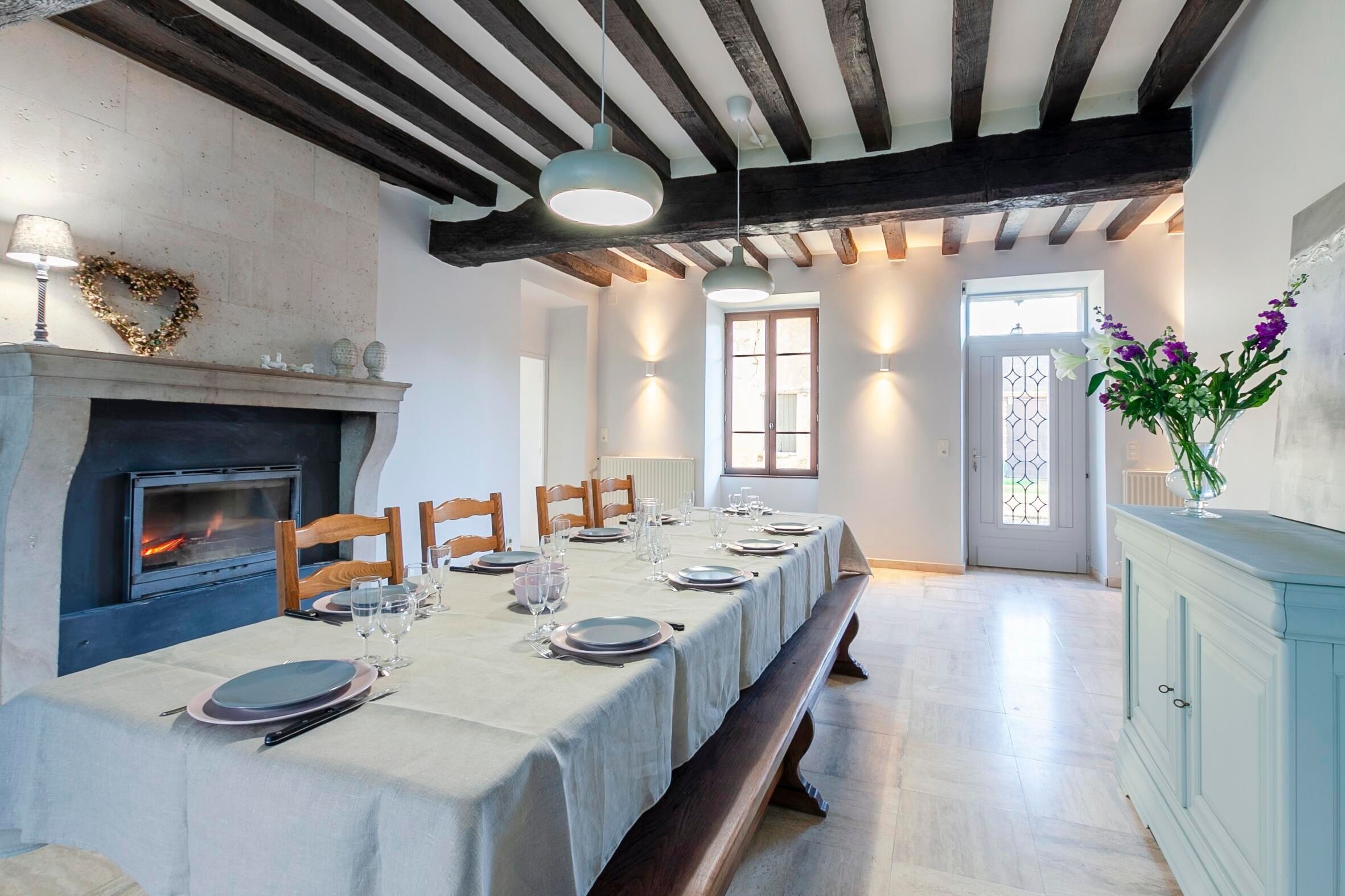 A dining area with a long table set for a meal, a fireplace, and a view through a window.