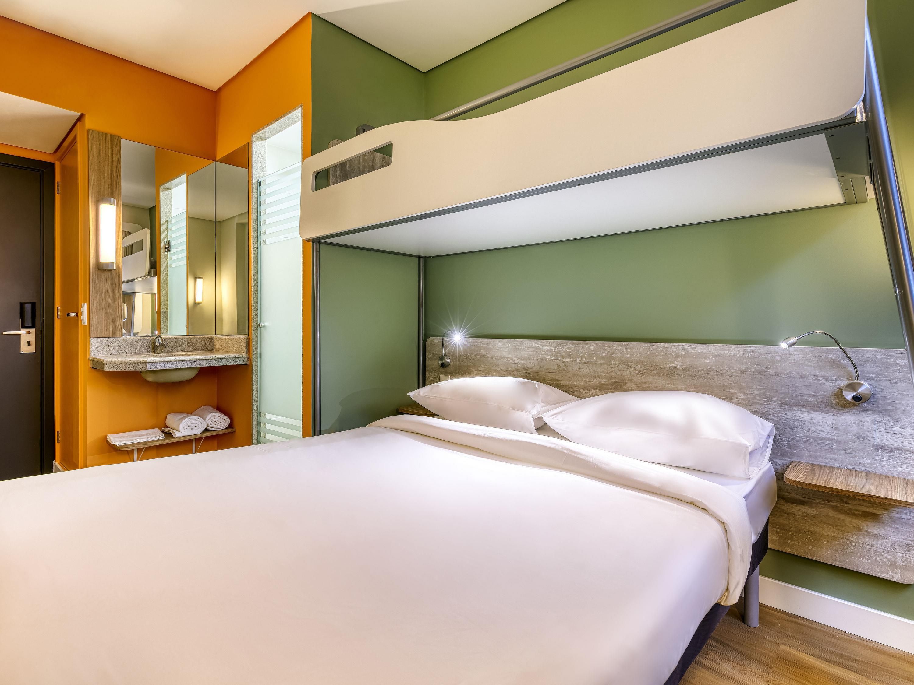 Una habitación de hotel moderna con una cama litera, un espejo grande, un lavamanos y una mesita de noche.