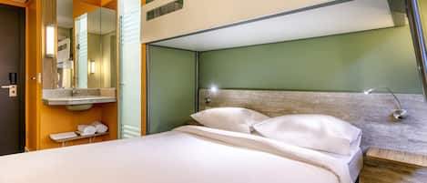 A modern hotel room with a bunk bed, a large mirror, a sink, and a bedside table.