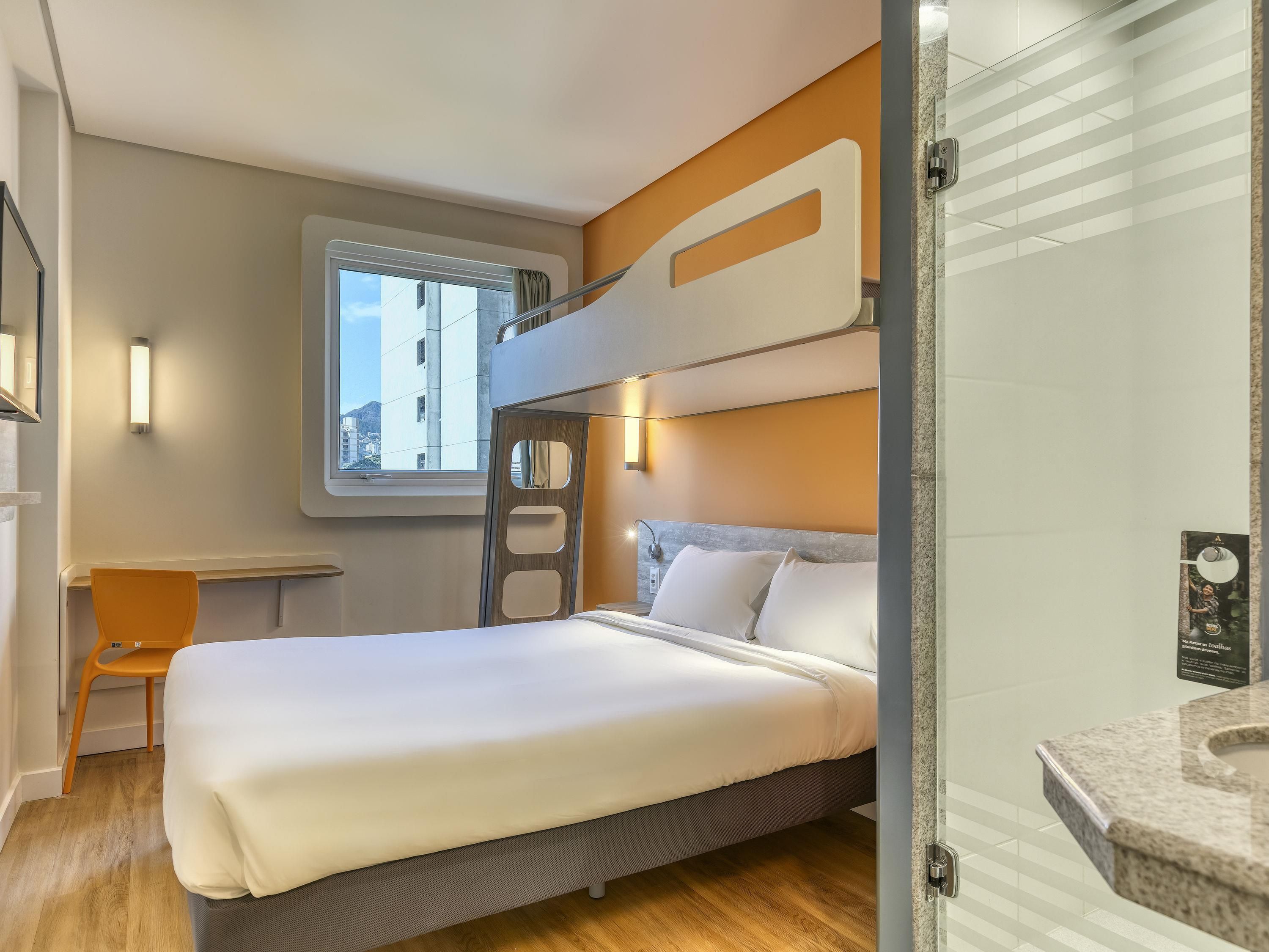 A hotel room with a bunk bed, a single bed, a window with a view of buildings, a small desk, and a bathroom with a granite countertop.