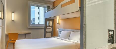 Quarto de hotel com beliche, uma cama de solteiro, janela com vista para edifícios, pequena escrivaninha e banheiro com bancada de granito.