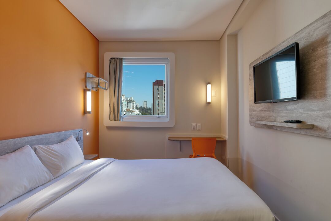 A hotel room with a bed, a TV mounted on the wall, a window with a view of buildings, and a small table with a chair.