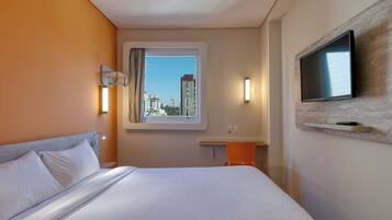 A hotel room with a bed, a TV mounted on the wall, a window with a view of buildings, and a small table with a chair.