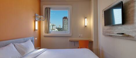 A hotel room with a bed, a TV mounted on the wall, a window with a view of buildings, and a small table with a chair.