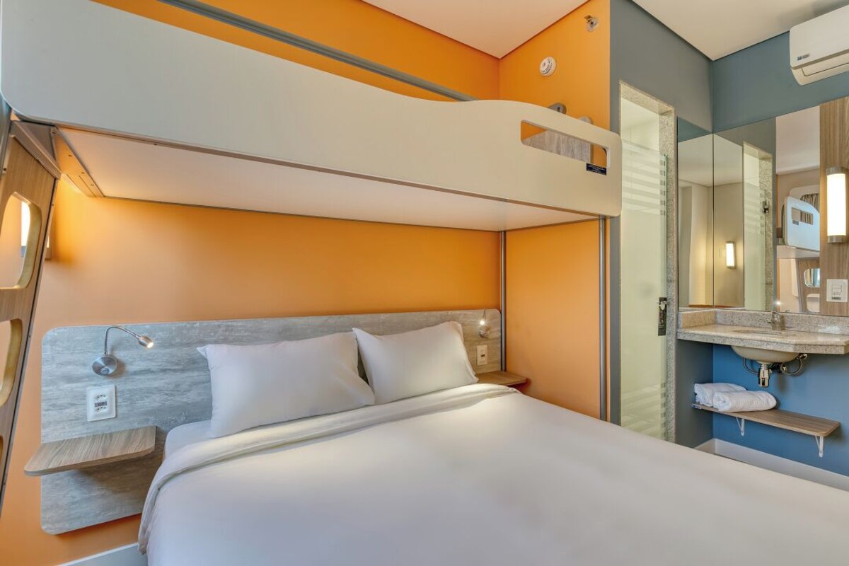 Une chambre d’hôtel avec un lit superposé, une table de chevet, un miroir et une salle de bain équipée d’un lavabo et d’un porte-serviettes.