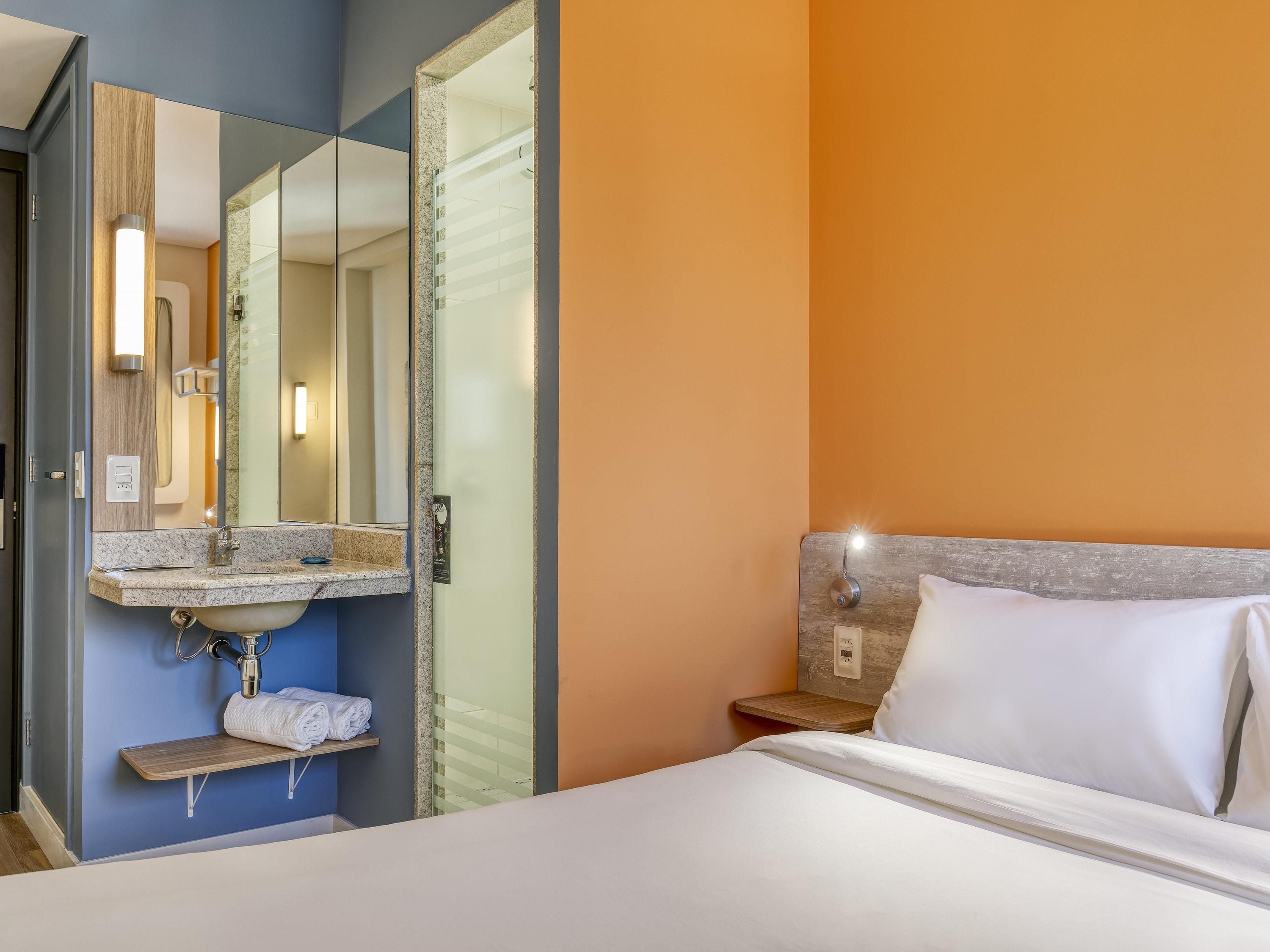 Una habitación de hotel con cama, baño con lavamanos y espejo, y una mesita de noche.