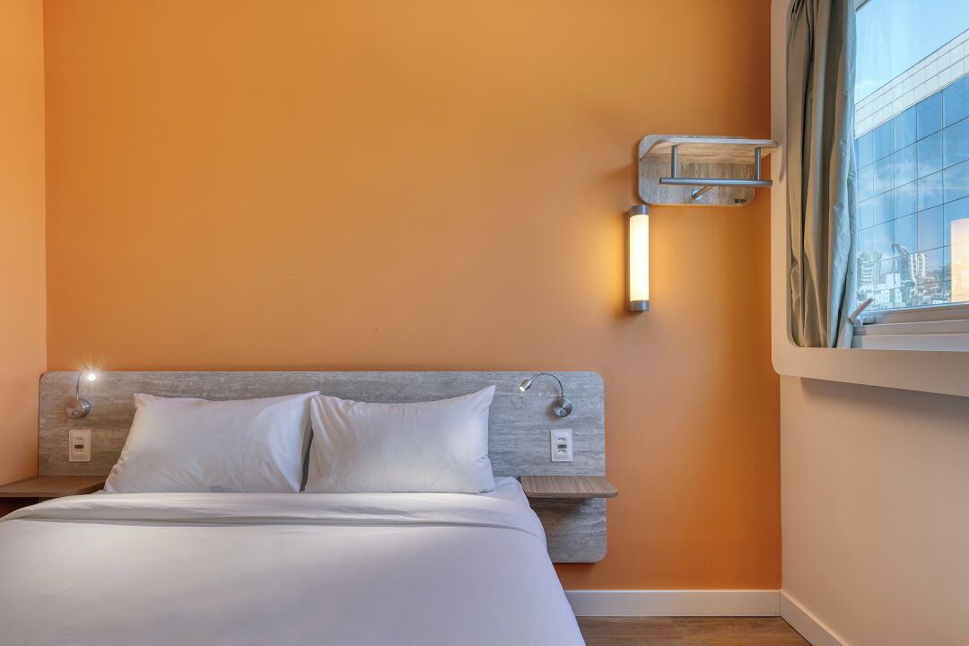 Quarto de hotel com cama, mesa de cabeceira, luminária na parede e janela com vista para edifícios.
