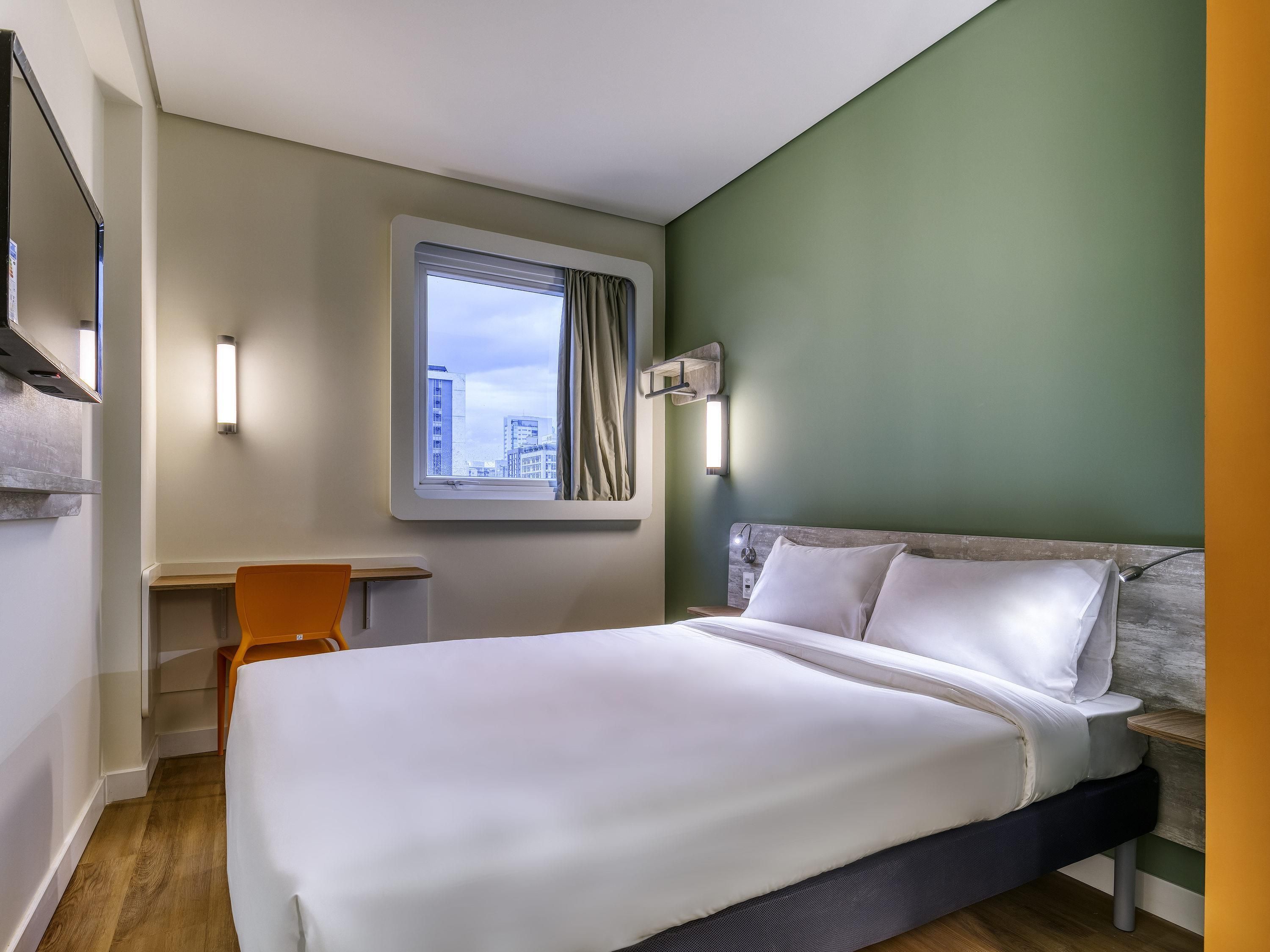 A hotel room with a bed, a desk, a chair, a window with a view of buildings, and a wall-mounted light fixture.