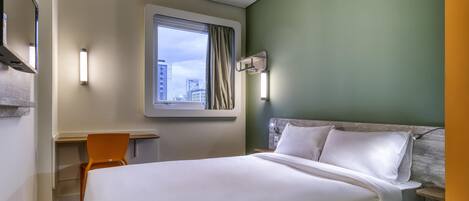 A hotel room with a bed, a desk, a chair, a window with a view of buildings, and a wall-mounted light fixture.