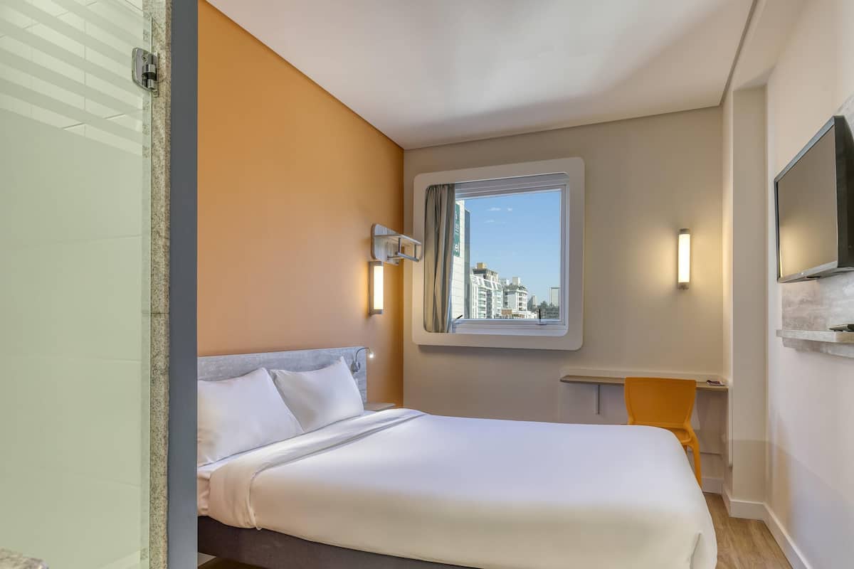 Habitación de hotel con cama, ventana con vistas a edificios, televisor de pantalla plana y una pequeña mesa.