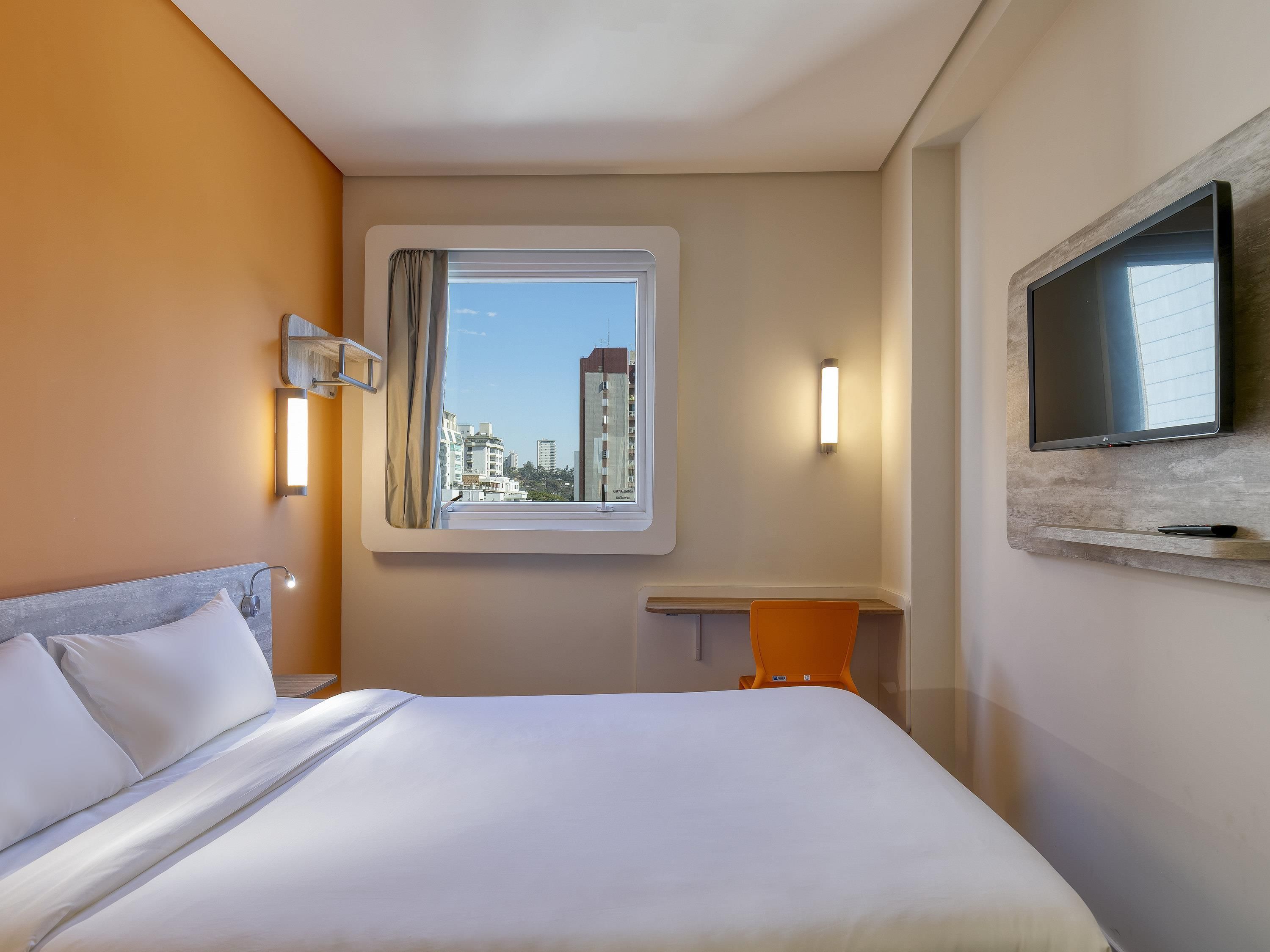 Quarto de hotel com cama, escrivaninha, televisão fixada na parede e janela com cortina.