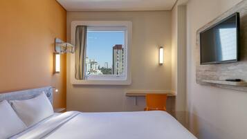 Quarto de hotel com cama, escrivaninha, televisão fixada na parede e janela com cortina.