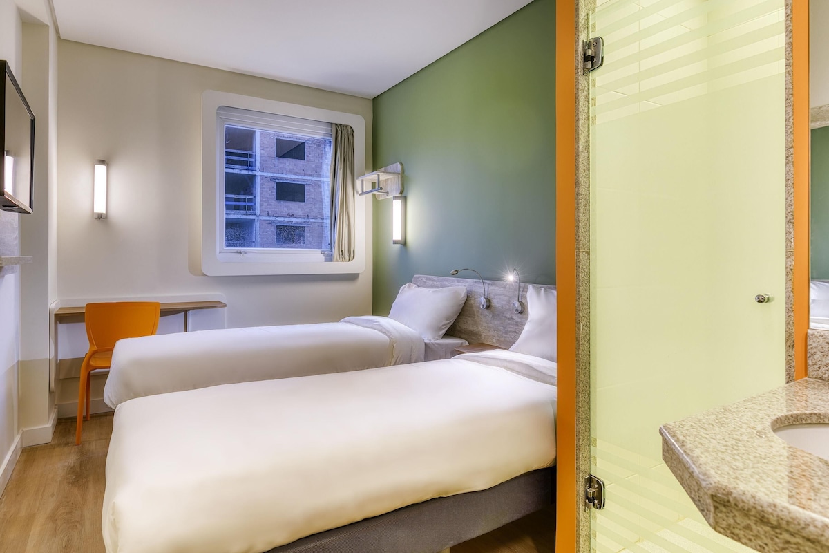 Une chambre d’hôtel avec deux lits, une petite table, une chaise, une fenêtre donnant sur un immeuble, et une salle de bain avec un lavabo et un miroir.