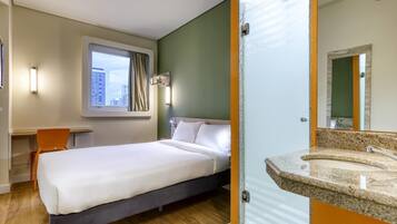 Quarto de hotel com cama, escrivaninha, janela com cortinas e banheiro com espelho e pia.