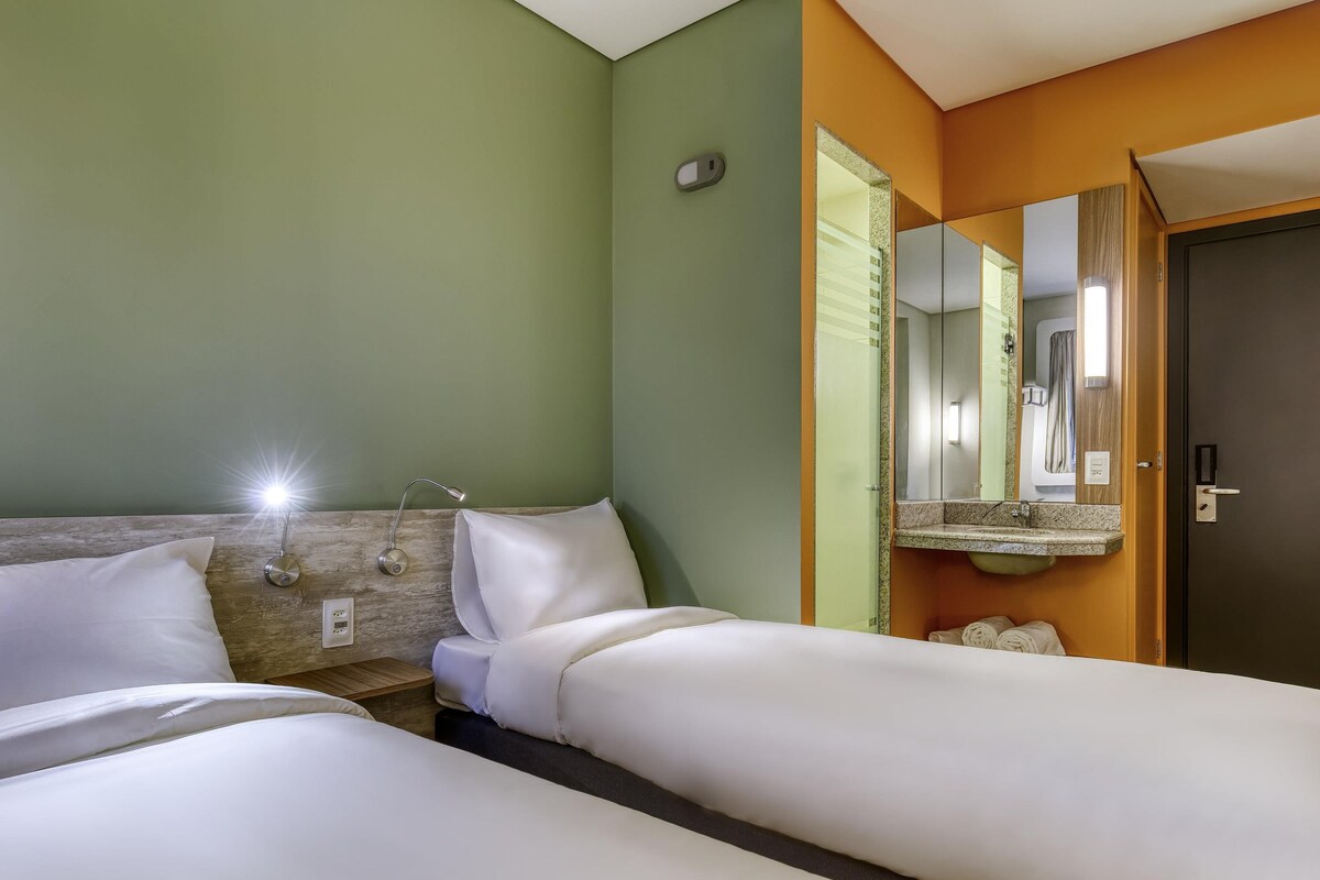 Ein Hotelzimmer mit zwei Betten, einem Nachttisch, einer Lampe, einem Spiegel und einer Tür.
