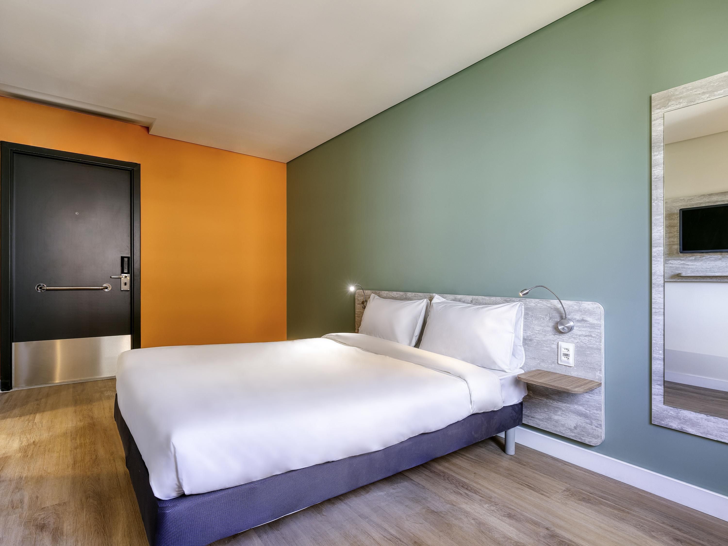 Quarto de hotel moderno com cama, mesa de cabeceira, televisão na parede e porta com puxador.