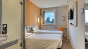 A hotel room with two beds, a window with a city view, a television mounted on the wall, and a small desk.