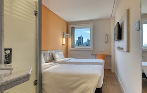 Ein Hotelzimmer mit zwei Betten, einem Fenster mit Stadtblick, einem an der Wand befestigten Fernseher und einem kleinen Schreibtisch.