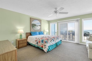 A bedroom with a bed, nightstands, a ceiling fan, a large window, and a view of the ocean.
