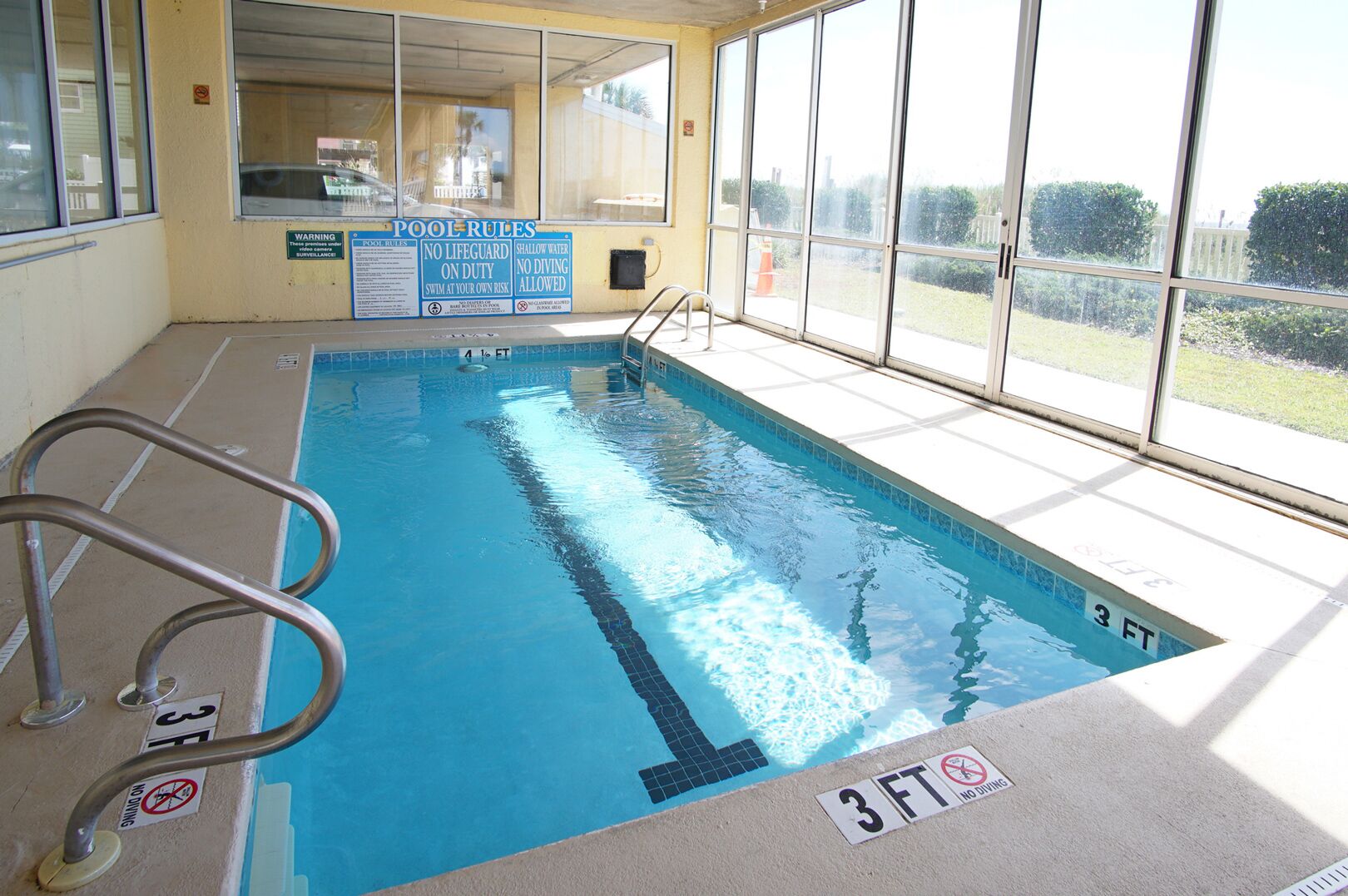 An indoor swimming pool with clear pool rules posted, a lifeguard chair, and a view of the outdoors through large windows.