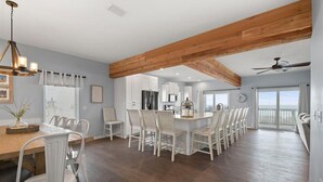 A spacious kitchen with wooden beams, a dining area with white chairs, and a view to the outside.