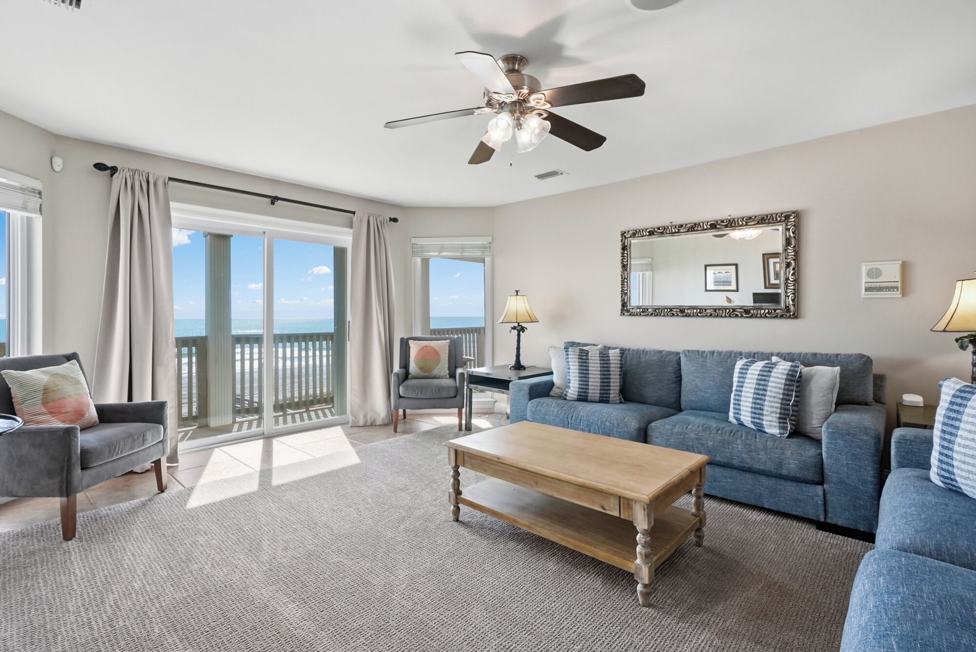 A spacious living room with a large window offering a view of the ocean, a ceiling fan, a blue sofa set, and a wooden coffee table.