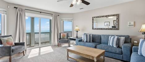 A spacious living room with a large window offering a view of the ocean, a ceiling fan, a blue sofa set, and a wooden coffee table.