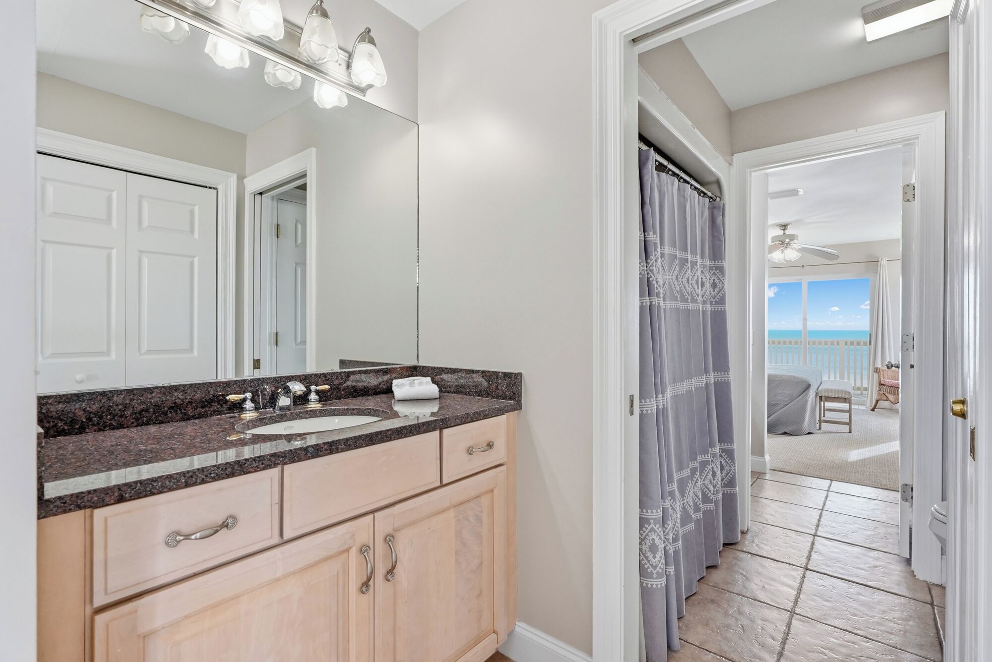 A bathroom with a granite countertop, a sink, and a mirror. There is a door leading to a balcony with a view of the ocean.