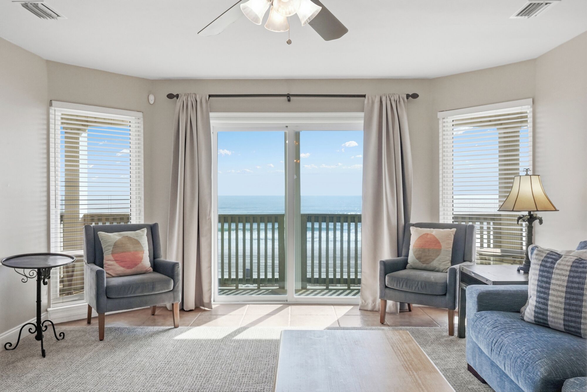 A room with two armchairs, a sofa, a small table, and a lamp. There is a view of the ocean through a glass door.